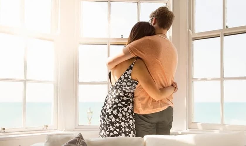 Casal jovem se abraça e olha pela janela da sala. Imagem ilustrativa. (Foto: Nick Doldin via Getty Images)